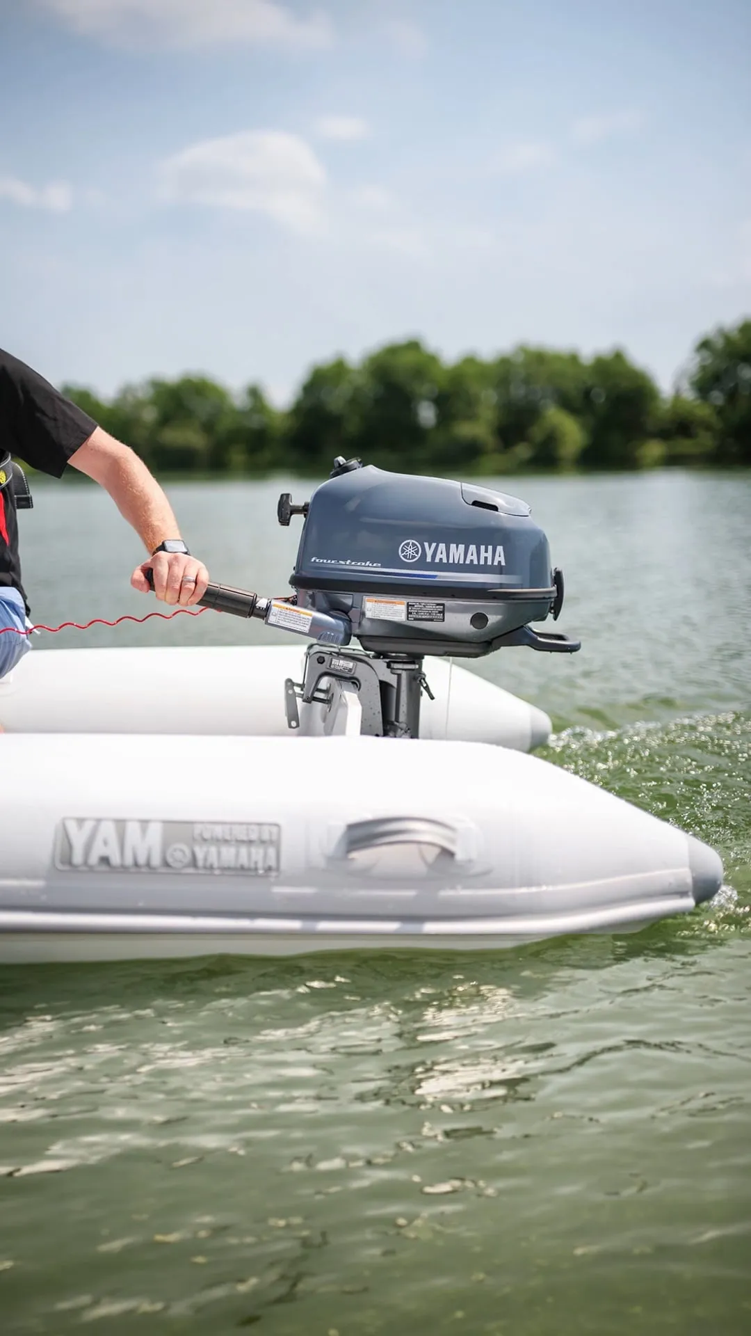 Person steering a small grey Yamaha four-stroke outboard by its tiller on a white YAM inflatable tender on a lake